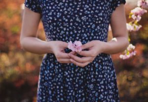 Eine Fraue, mit blau-weiß geblümten Kleid, hält ein zartes rosa Blümchen in den Händen