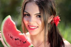 Eine junge Frau mit braunem Haar und rotem Haarband hält lächelnd eine Wassermelonenspalte in der Hand.