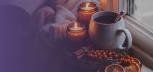 Das Bild zeigt ein warmes, gemütliches Herbst- und Wintermotiv. Auf einem Holzbrett an der Fensterbank stehen eine Keramiktasse mit einem Heißgetränk sowie zwei brennende Kerzen in braunen Apothekergläsern. Davor liegen Zimtstangen, ein Zweig mit orangefarbenen Sanddornbeeren und getrocknete Orangenscheiben. Im Hintergrund befinden sich eine weiße Wolldecke und ein geöffnetes Buch. Die linke Seite des Bildes ist mit einem lila Verlauf schattiert, um Text einzublenden.