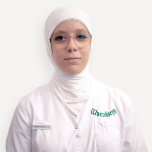 Amna Alibegovic, Drogistin in Ausbildung bei Wurzelsepp, lächelt in die Kamera. Sie trägt ein weißes Kopftuch, eine Brille und einen Arbeitskittel mit Namensschild und Firmenlogo.
