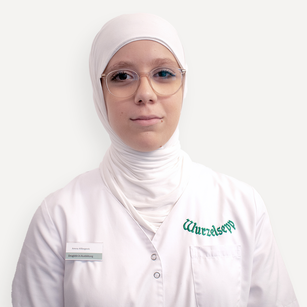Amna Alibegovic, Drogistin in Ausbildung bei Wurzelsepp, lächelt in die Kamera. Sie trägt ein weißes Kopftuch, eine Brille und einen Arbeitskittel mit Namensschild und Firmenlogo.